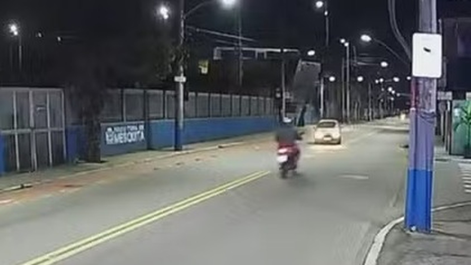 Rua noturna com moto, carro, postes de iluminação e muro azul com letreiro