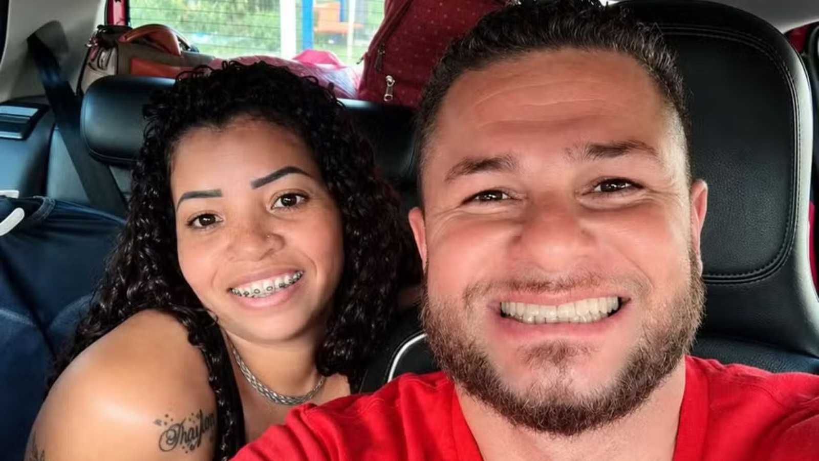 Casal sorridente em selfie no carro, homem com barba e mulher com aparelho dental