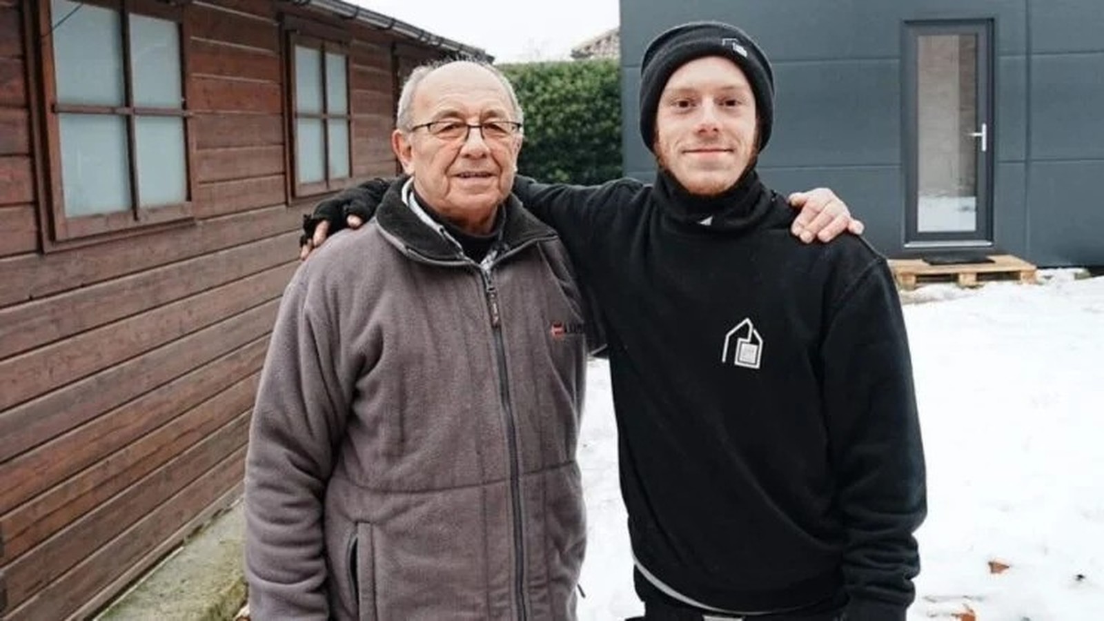 Homem idoso com óculos e homem jovem em quintal nevado, braços um sobre o outro