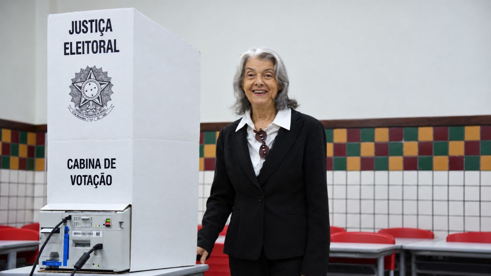 Mulher sorridente ao lado de cabina de votação eletrônica. Texto: JUSTIÇA ELEITORAL e CABINA DE VOTAÇÃO