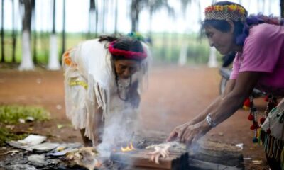 Duas mulheres indígenas manuseiam alimentos em fogo aberto com fumaça, árvores desfocadas no fundo