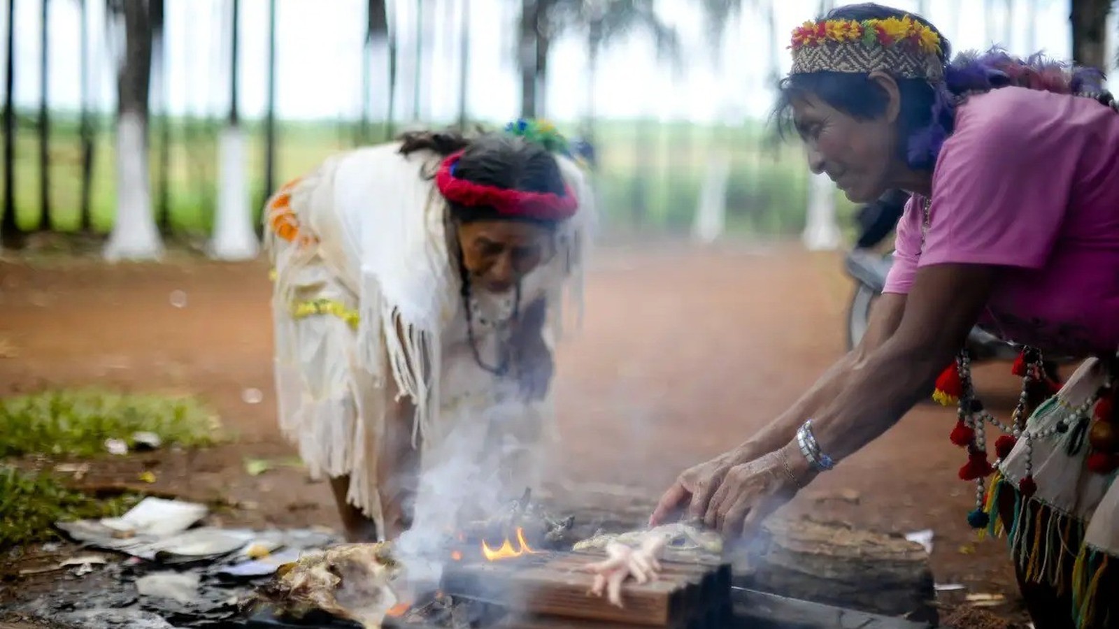 Duas mulheres indígenas manuseiam alimentos em fogo aberto com fumaça, árvores desfocadas no fundo