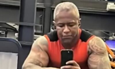 Homem musculoso de pele escura, cabelo curto claro, camisa vermelha, tirando selfie com celular