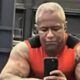 Homem musculoso de pele escura, cabelo curto claro, camisa vermelha, tirando selfie com celular