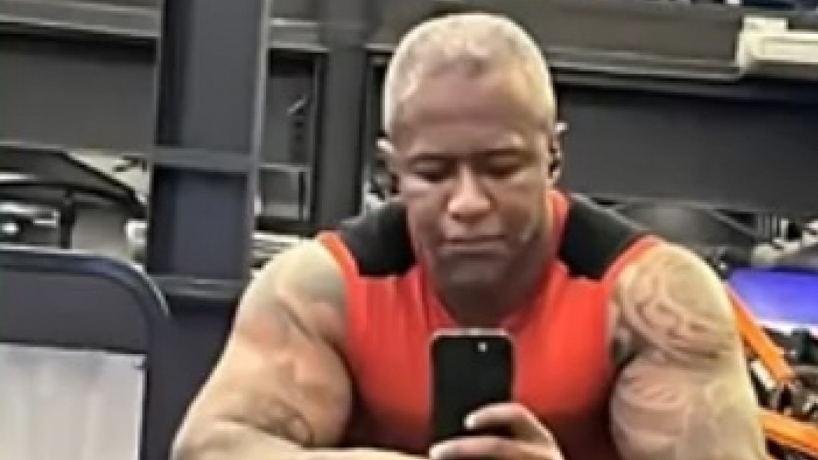 Homem musculoso de pele escura, cabelo curto claro, camisa vermelha, tirando selfie com celular
