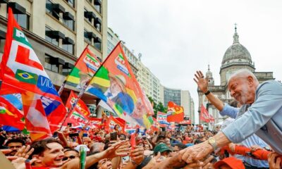 Lula sorrindo, acenando para multidão em comício, com bandeiras vermelhas "LULA" e "VICL"