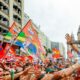 Lula sorrindo, acenando para multidão em comício, com bandeiras vermelhas "LULA" e "VICL"