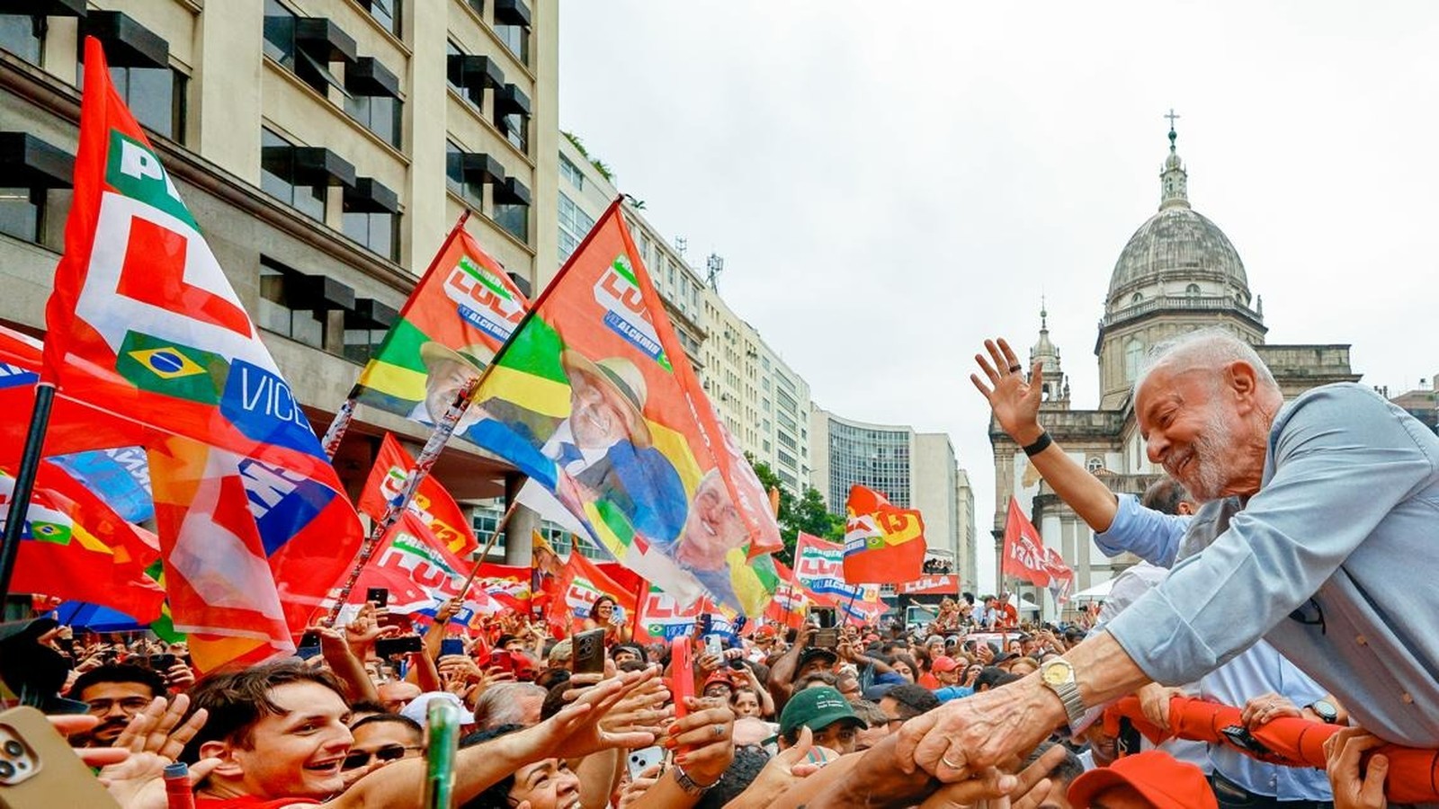 Lula sorrindo, acenando para multidão em comício, com bandeiras vermelhas "LULA" e "VICL"