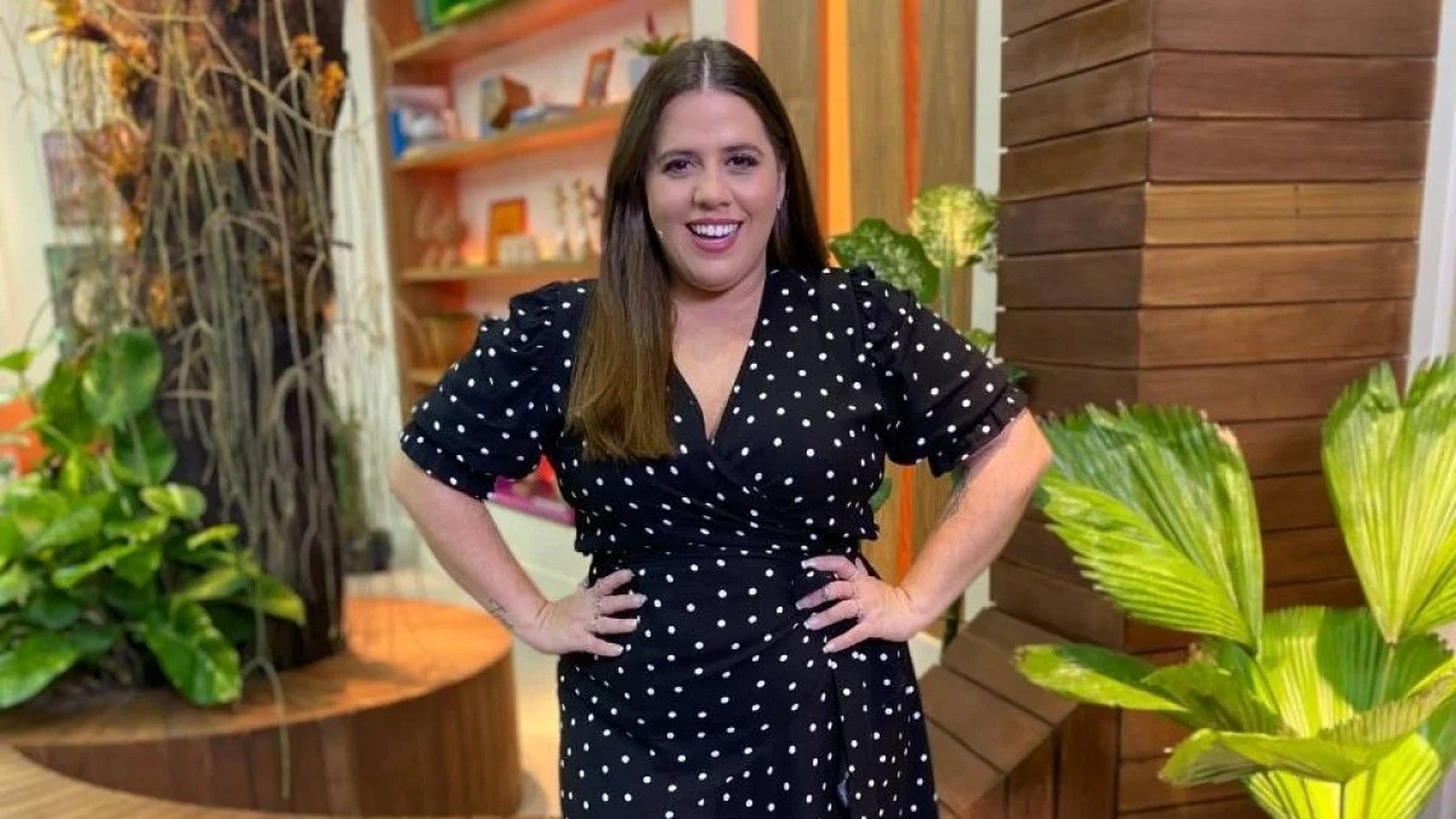 Tati Machado sorrindo, vestido preto de bolinhas brancas, mãos na cintura.