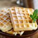 Três waffles dourados com açúcar de confeiteiro e um ramo de hortelã em prato de madeira