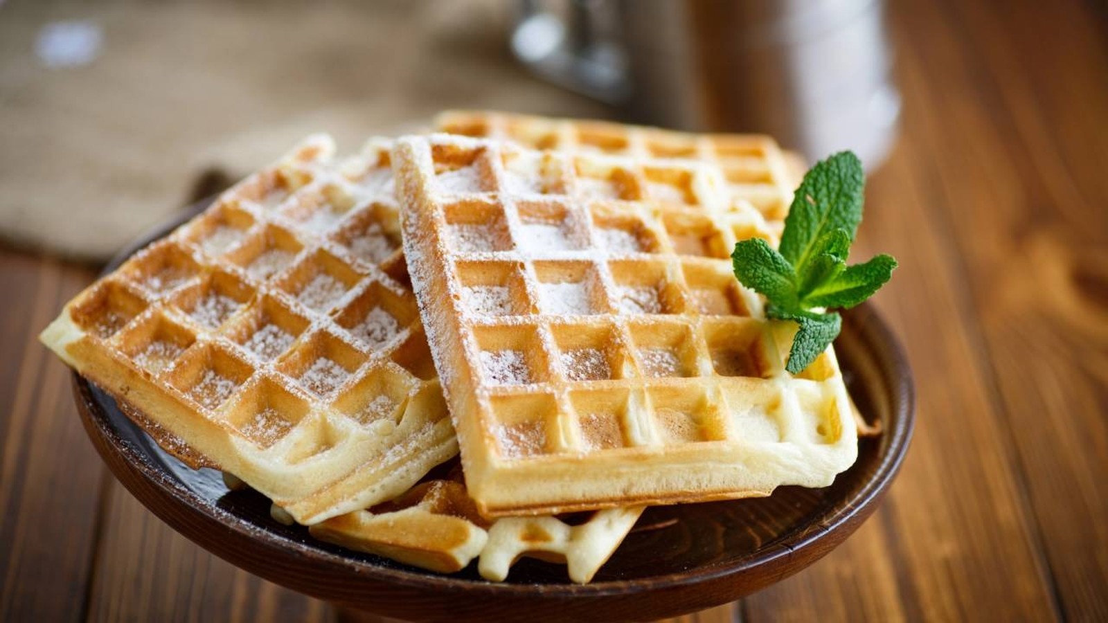 Três waffles dourados com açúcar de confeiteiro e um ramo de hortelã em prato de madeira