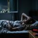 Mulher deitada na cama acordada de noite, com mão na testa, e relógio digital marcando 03:47