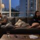 Homem e mulher sentados distantes em um sofá cinza na sala, com expressões tristes