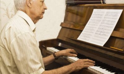 Homem idoso de perfil tocando piano de madeira com partitura e mãos sobre as teclas