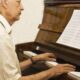 Homem idoso de perfil tocando piano de madeira com partitura e mãos sobre as teclas