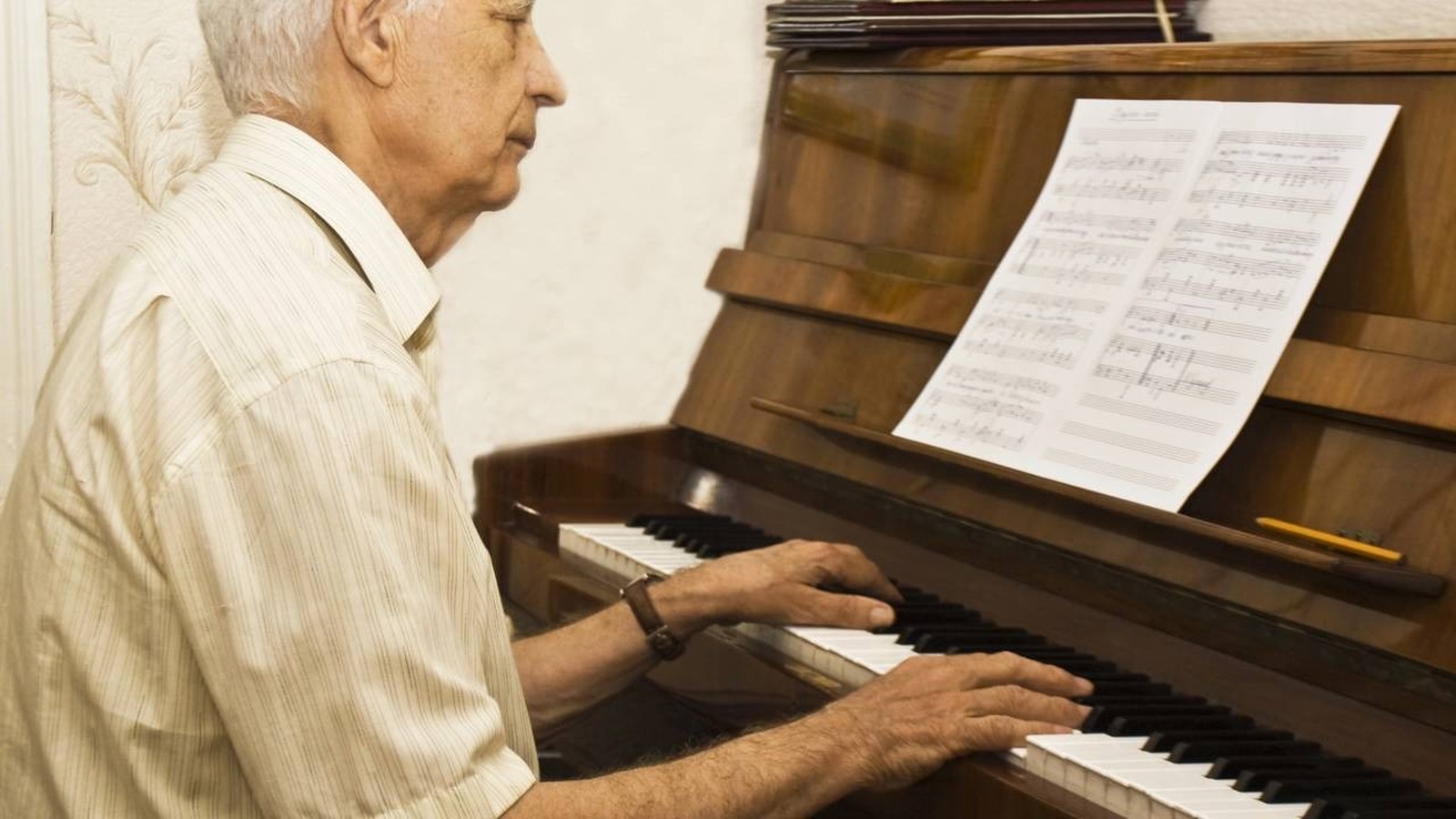Homem idoso de perfil tocando piano de madeira com partitura e mãos sobre as teclas