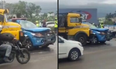 Caminhão amarelo colidiu com viatura policial azul danificada. Cena de acidente em rua molhada, com pessoas